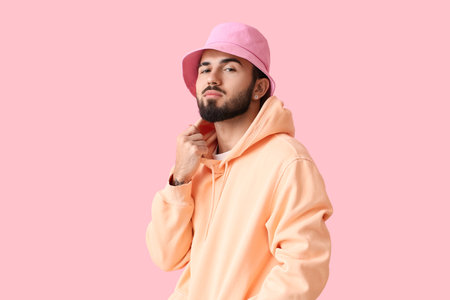 Fashionable bearded man wearing hoodie and hat on pink background