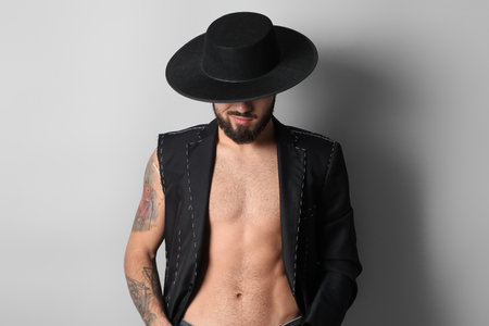 Young bearded man in half-finished jacket and stylish hat on light background