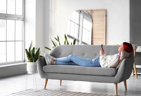 Pretty young woman with unusual hair and phone lying on sofa at homeの写真素材