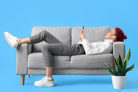 Pretty young woman with unusual hair and phone lying on sofa against blue backgroundの写真素材