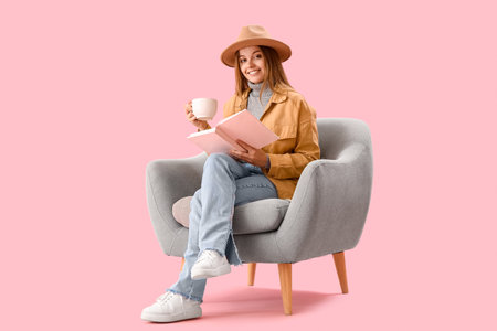 Young woman with cup of hot cocoa reading book in armchair on pink backgroundの写真素材
