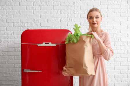 Woman with bag of fresh products near stylish red fridge on white brick wall backgroundの写真素材