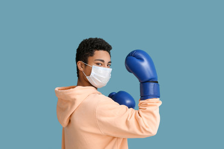Young African-American man in medical mask with boxing gloves on blue background. immunity conceptの写真素材