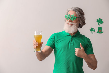 Senior man in eyeglasses with glass of beer showing thumb-up on light background. St. Patrick's Day celebrationの写真素材