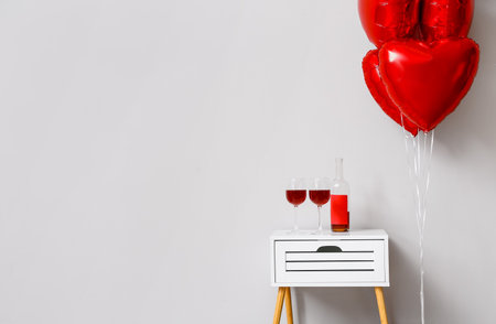 Beautiful heart-shaped balloons with glasses and bottle of wine for Valentine's Day celebration near light wallの写真素材