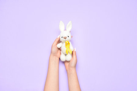 Woman holding toy rabbit with golden awareness ribbon on violet background, closeup. International Childhood Cancer Dayの写真素材
