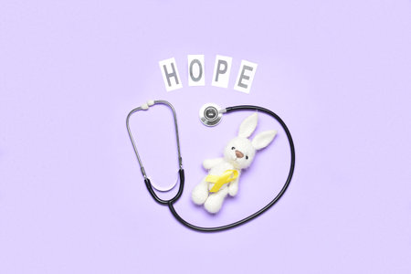 Golden awareness ribbon, stethoscope, toy rabbit and word HOPE on violet background. International...