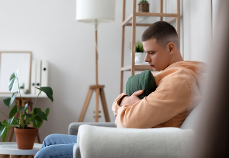 Worried young man with pillow sitting on sofa at homeの写真素材