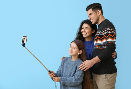 Little girl and her parents in warm sweaters taking selfie on blue background