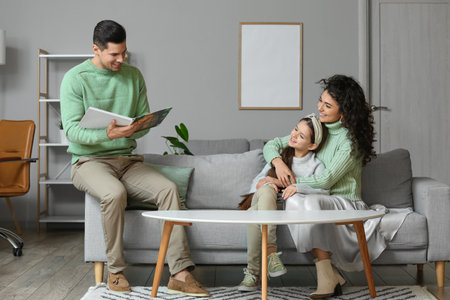 Happy young man reading book to his wife and little daughter at homeの写真素材