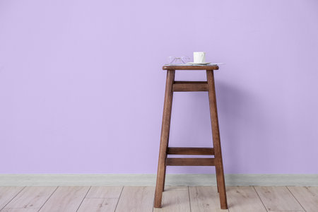 Wooden bar stool with cup of coffee and eyeglasses near violet wall in roomの写真素材