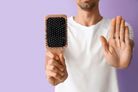 Young man holding brush with fallen down hair and showing stop gesture on lilac background