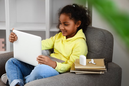 Little African-American girl studying with online tutor in armchair at home