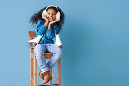 Little African-American girl with ice skates sitting in chair on blue backgroundの写真素材