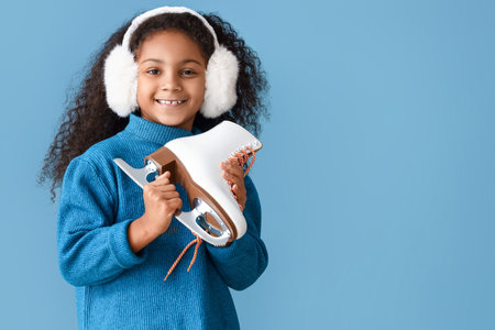 Little African-American girl in earmuffs with ice skates on blue backgroundの写真素材