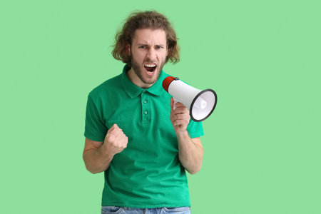 Angry young man shouting into megaphone on green background