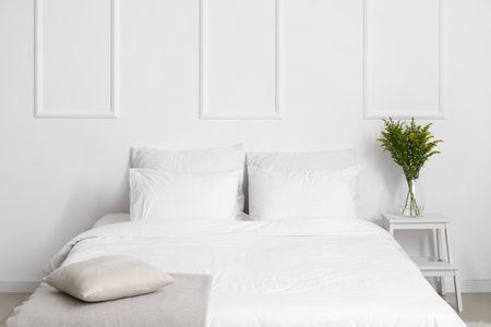 Comfortable bed and vase with flowers on stepladder near white wallの写真素材