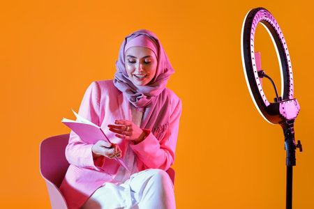 Muslim female blogger with notebook recording video on yellow background