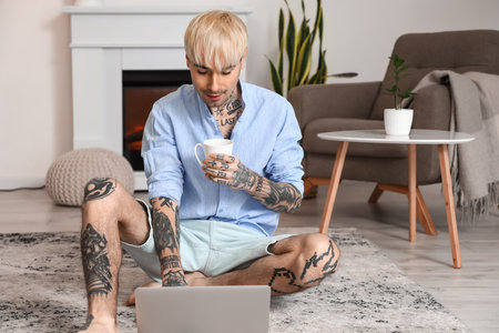 Young tattooed man with cup of coffee using laptop on floor at homeの写真素材