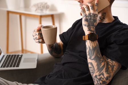 Young tattooed man with cup of coffee and laptop talking by mobile phone at homeの写真素材