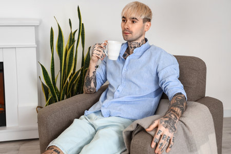 Young tattooed man with cup of coffee sitting in armchair at homeの写真素材