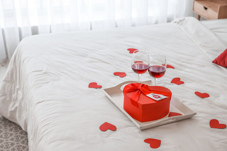 Tray with gift for Valentine's Day and glasses of red wine on bedの写真素材