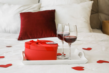 Tray with gift for Valentine's Day and glasses of red wine on bedの写真素材