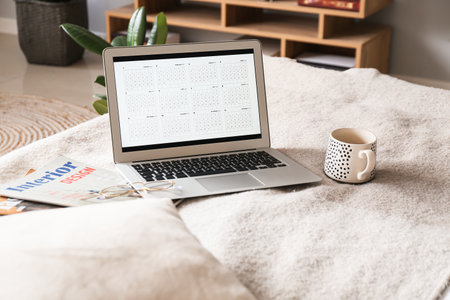 Laptop with digital calendar, eyeglasses, magazines and cup on bed in roomの写真素材