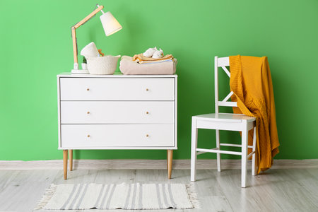 Chest of drawers with baby clothes and chair with knitted sweater near color wall in room interiorの写真素材