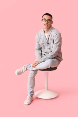 Handsome young Asian man in knitted sweater sitting on chair against pink backgroundの写真素材