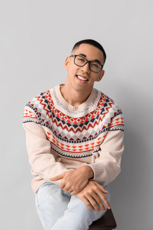 Handsome young Asian man in knitted sweater sitting on chair against gray backgroundの写真素材