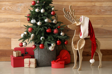 Decorative reindeer with Santa hat, Christmas tree and presents near wooden wallの写真素材