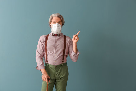 Senior man in medical mask with walking stick pointing at something on gray backgroundの写真素材