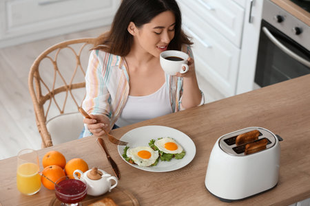 Beautiful young Asian woman drinking coffee and eating tasty toasts with fried eggs in kitchenの写真素材
