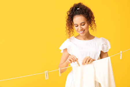 Young African-American woman hanging clean laundry with wooden clothespin on yellow backgroundの写真素材