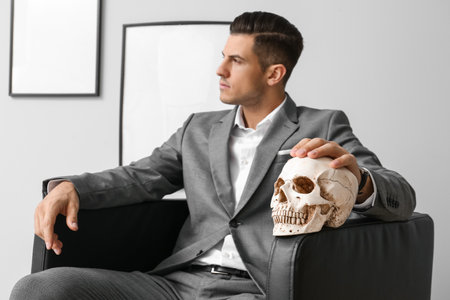 Fashionable handsome man sitting in armchair with skull and looking aside near gray wallの写真素材