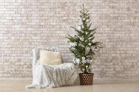 Beautiful Christmas tree in pot decorated with snowflakes, balls and comfortable armchair near beige brick wallの写真素材