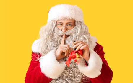 Santa Claus with Christmas bell showing silence gesture on yellow backgroundの写真素材