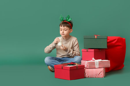 Funny little boy with Christmas gifts drinking milk on color background