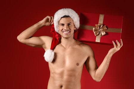 Sexy young man in Santa hat and with Christmas gift on color backgroundの写真素材