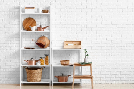 Modern shelf units with kitchenware and stand near white brick wall