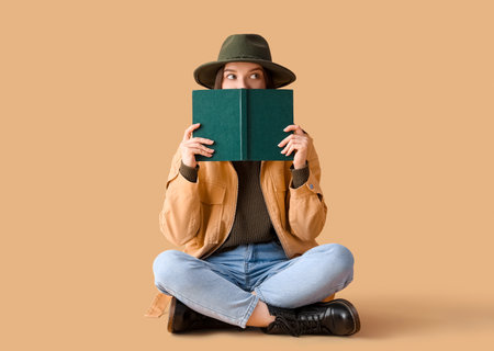 Young woman in hat reading book on beige background