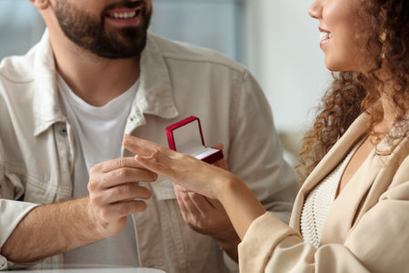 Young man putting engagement ring on his girlfriend's finger at homeの写真素材