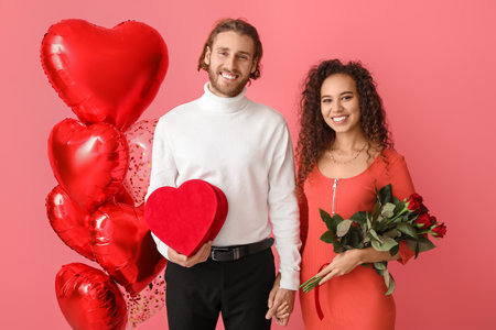 Happy young couple with balloons, gift and flowers on color background. Valentine's Day celebration