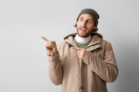 Handsome young man in winter clothes pointing at something on light background