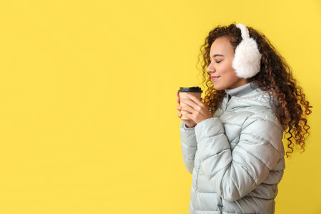 Beautiful African-American woman in winter clothes and with cup of coffee on color backgroundの写真素材