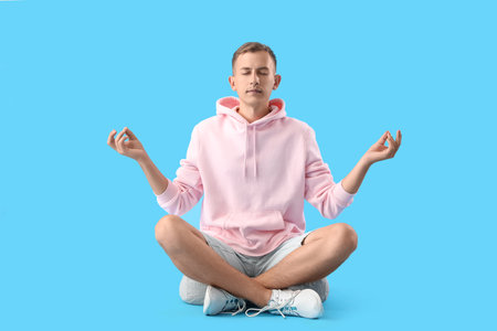 Young man in pink hoodie meditating on blue backgroundの写真素材