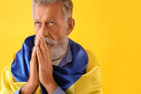 Mature man with flag of Ukraine praying on yellow background, closeupの写真素材