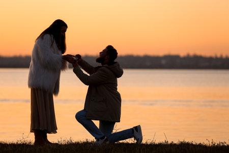 Young man proposing to his girlfriend near river at sunsetの写真素材