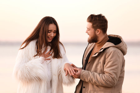 Young man putting engagement ring on finger of his fiancee near riverの写真素材